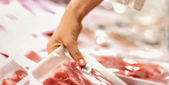 A person's hand reaches for a package of raw meat in a grocery store's refrigerated section, mindful of pathogen testing. The meat is in a white tray wrapped with clear plastic, with other trays blurred in the background. - Matrix Sciences